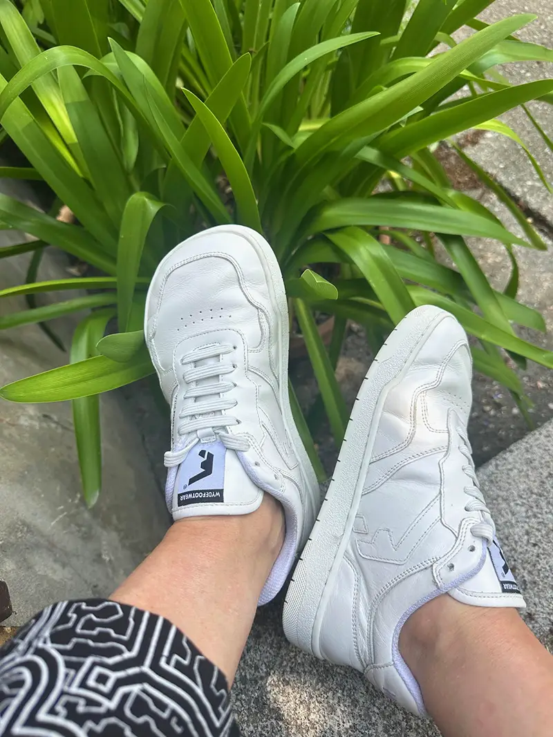 White sneakers worn outdoors, feet resting near green plants