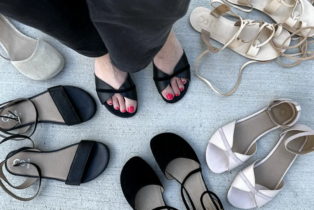 Assorted barefoot dress shoes and sandals with wide toe boxes in neutral colors, displayed on light concrete, including black, beige, and blush styles.