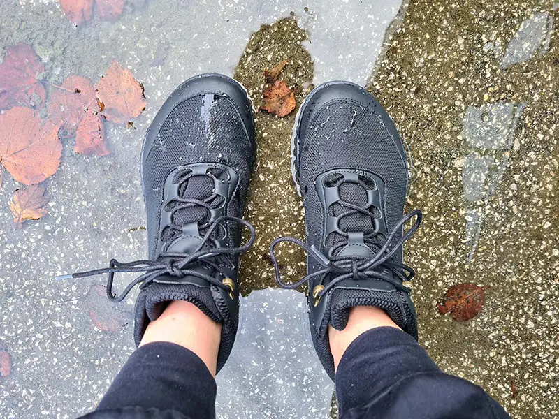 Black waterproof barefoot boots standing on wet pavement with raindrops on the upper.